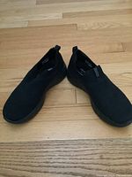 View of the Skechers black slip-on shoes from an angle showing both shoes placed on a wooden floor. Visible knit texture on upper, pull tabs present.