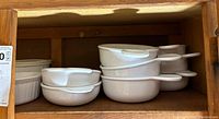 White ceramic round baking dishes with handles stacked inside each other inside wooden shelf.