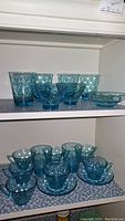 Full set of blue Hazel Atlas Capri Dot glassware displayed on two shelves. Top shelf has mixed glasses and bowls; bottom shelf has cups with saucers and smaller cups.