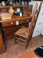 Photo showing the wood writing desk with two front drawers and shelves underneath. Partial view of wood ladder back chair with rush seat in front of desk.
