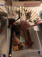 Two decorative hand fans inside a plastic container with various small wooden items including nesting dolls and a dog figurine.