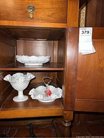 Three white milk glass hobnail serving bowls placed on wooden shelves inside a cabinet, including a footed bowl and two shallow bowls