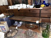 Photo showing three tier wood shelves with black metal frame. Upper and middle shelves hold various household items. Visible scratches and wear on wood surfaces.