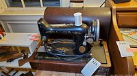 Black Singer portable electric sewing machine with gold floral decals, mounted on a wooden base with power cord and foot pedal. Includes spool of white thread on spool pin.