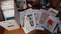 Wide view of a box and loose stack of vintage brochures and paper ephemera projects, showing some titles and designs.