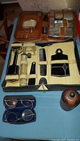 Overall shot of two vintage men's grooming kits displayed open showing brushes, combs, scissors, razors, and toiletry containers, two pairs of spectacles in a blue case, and a wooden container.