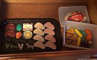 Full image showing a black tray of plastic play food pieces including bacon, vegetables, salmon, steak, and metal cookie cutters shaped like farm animals, plus plastic storage containers holding additional play food items such as pie slices and vegetables.