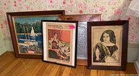Photo showing four framed wall art pieces grouped on floor, including sailboat painting, vintage magazine cover, another wooden framed artwork, and a vintage-style portrait of a girl.