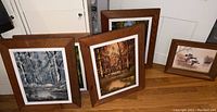 Four framed prints arranged on floor showing nature scenes including winter birch trees, autumn pond, and ducks on water