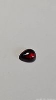 Close-up image of a pear-shaped cherry red garnet gemstone with a glossy polished surface against a plain background.