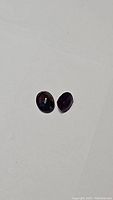 Photo of two oval faceted sapphire gemstones showing dark pink, blue, and purple colors on a white background.