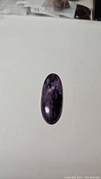 Oval purple charoite cabochon gemstone displaying its deep purple color with black inclusions placed on a white surface under general ambient lighting.