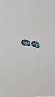 Pair of oval cut paraiba blue apatite gemstones, 6x4mm each, placed on a plain surface showing their vibrant blue color and clarity.