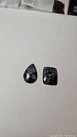 Photo showing two polished cabochons of snowflake obsidian on white background, one pear-shaped and one cushion-shaped.