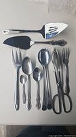 Top-down view of 11 serving utensils arranged on white background showing variety of pie and cake servers, spoons, forks, salad servers, and tongs.
