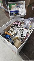Box with assorted handyman items including cables, clamp with wooden handle, twine spool, scissors, plastic containers, and various small hardware pieces.