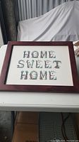 Full view of the framed wall art showing the phrase "Home Sweet Home" in decorative floral lettering within a medium brown wooden frame.