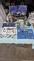 Full view of all silver plate pieces laid out on table showing various items including coffee pot, trays, casseroles, and assorted cutlery.