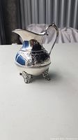 Silver plated engraved creamer with floral design and scalloped details standing on four decorative feet, shown on white surface with background showing vertical blinds.