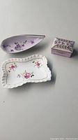 Three china trinket trays/boxes visible: white floral tray, purple teardrop tray with chip, and purple lidded box with gold accents