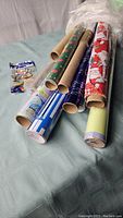 Rolls of assorted wrapping paper featuring different patterns including Christmas Santa Claus, blue snowflakes, green hearts, kraft paper, and striped designs, laid out on a fabric surface.