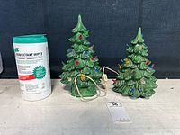 Both ceramic Christmas trees positioned side by side next to a container of disinfectant wipes for size reference. One tree has a green base and power cord, the other does not.