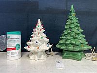 Both ceramic Christmas trees together next to disinfectant wipes for scale. Shows medium white tree on left with multicolored bulbs and star-shaped base, and large green tree on right also with bulbs and base. Electric cords are visible.