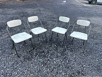 Four vintage mid century metal folding chairs with white padded seats and backrests arranged outdoors on a gravel surface.