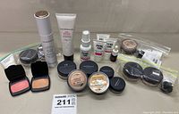 Group of Bare Minerals beauty products including lotions, foundation powders, primer tubes, blush compacts, serum bottle, and additional makeup containers all arranged on a grey surface.