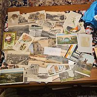 Photo showing a spread of various vintage postcards on a table, featuring black and white images of city buildings, bridges, and some illustrated greeting cards.