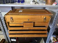 Front of the wooden workers chest with the front panel removed showing three drawers and one small central compartment. The chest is light brown wood with metal clasps on either side and a lock hole without key.