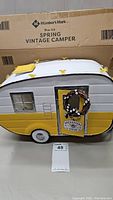 Front side view of the yellow and white vintage camper decoration showing the door with wreath and sign, and one window.
