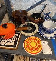 Grouping of vintage kitchen items including wooden salad bowls, skillet with wooden handle, bright orange teapot, blue and white ceramic pitcher and cup, decorative plate, and coaster.