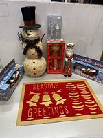 Wide view showing a snowman decoration, boxed angel, ceramic Santa, two boxed Flying Santa Sleigh toys and a seasonal door mat laid flat.
