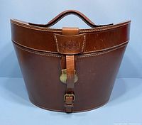 Front view of brown leather top hat box with handle, showing buckle, strap, embossed emblem, and white stitching.