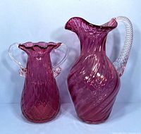 Front view of cranberry glass pitcher with twisted clear handle and matching cranberry vase with two clear handles and ruffled top.