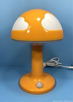 Front view of orange plastic lamp with white cloud shapes on shade, push-button switch visible on base, AC cord looped behind