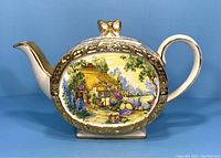 Front angle showing pastoral scene with cottage, flowers, and lady. Gold trim and bow detail on lid visible.