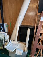 Large and two smaller rolls of natural off-white artist canvas leaning against a wood paneled wall, with pre-cut canvas pieces in a plastic bin below