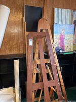 Five wooden artist easels of various sizes and a wood framed chalkboard displayed against a wood paneled wall with paintings nearby.