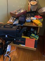Photo showing a table with photographic equipment including Minolta bag, film storage album, Kodak boxes, flash units, and camera accessories stacked on and around a wooden cabinet with shelves and drawer.