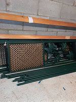 Four green metal hinged garden privacy screen panels stacked showing decorative leaf motif and woven lattice insert panels with wood posts on top.