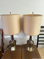 Pair of two matching 34 inch tall table lamps with beige fabric drum shades, round brass finials, and bases with brass and brown textured sections shown on wooden table between wooden chairs.