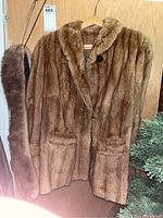 Full front view of brown mink fur coat hanging on wooden hanger, showing two front pockets and a fur collar. Matching fur stole/scarf hanging beside coat.