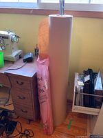 Large roll of brown craft paper standing on the floor next to desk, partly on a metal spindle. A partial roll of clear plastic sheeting is stored inside a pink plastic bag beside it.