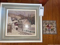 Photo shows framed rug hooking winter scene artwork with wood frame and hooked wool coaster with floral design to the side.