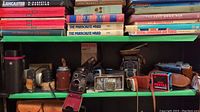 Shelf displaying multiple vintage cameras, leather cases, lens cases and boxes including Pentax Zoom 90 signage