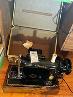 Singer vintage sewing machine with gold decal details, spool of white thread attached, and electrical cord connected, displayed alongside its open hard carrying case with brown interior.
