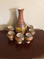 Front view of Peruvian hand painted clay carafe surrounded by six matching cups on a plain surface against a neutral wall.