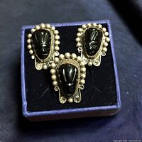 Set of one ring and pair of earrings displayed on black velvet in blue box. Each features black carved onyx face stone with silver beaded and scroll detailing.